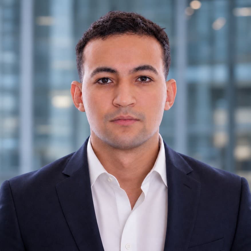 Will Bayoud in a navy jacket and white shirt, against a blue-grey architectural background